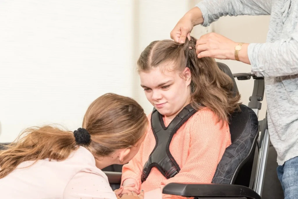 A disabled girl sitting in her wheelchair while her support worker holds her hand and fixes her hair